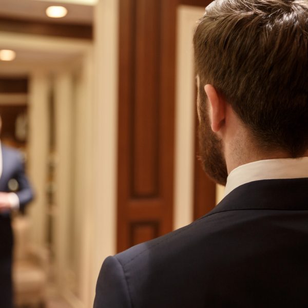 Man looking in mirror wearing suit Close up shot of back of bearded man wearing suit and looking at himself in mirror in wood dressing room